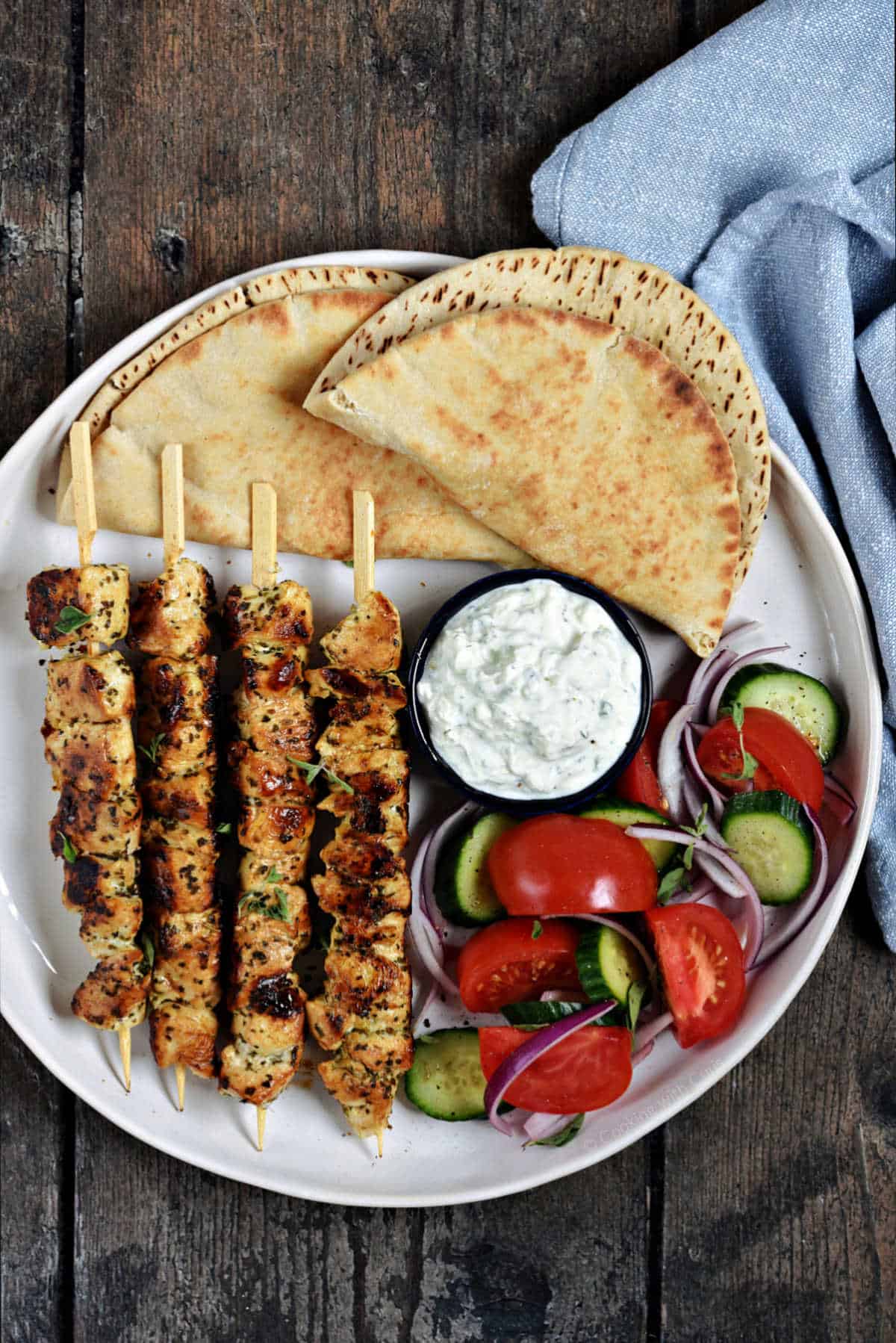 Greek Chicken Souvlaki served on a platter with warm pita, tzatziki, sliced tomatoes, red onion, and fresh parsley garnish