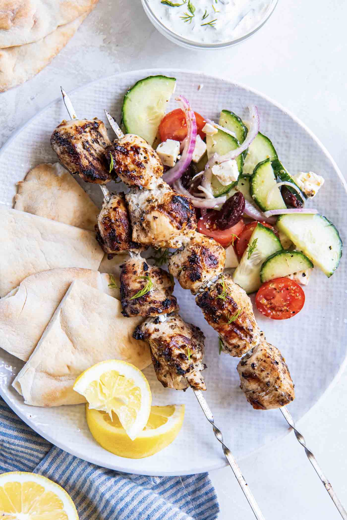 Close-up of Greek Chicken Souvlaki skewers served in warm pita with tzatziki, diced tomatoes, red onion, and fresh herbs on a rustic wooden board