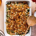 A golden-topped green bean casserole in a white ceramic baking dish, garnished with extra crispy fried onions, shot from a slight angle on a rustic wood Thanksgiving table