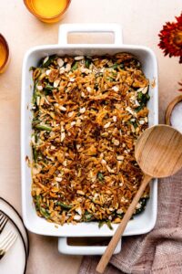 Green Bean Casserole — The Thanksgiving Standard Made From Scratch 13 A golden-topped green bean casserole in a white ceramic baking dish, garnished with extra crispy fried onions, shot from a slight angle on a rustic wood Thanksgiving table