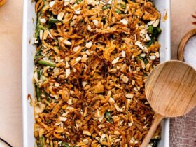 A golden-topped green bean casserole in a white ceramic baking dish, garnished with extra crispy fried onions, shot from a slight angle on a rustic wood Thanksgiving table