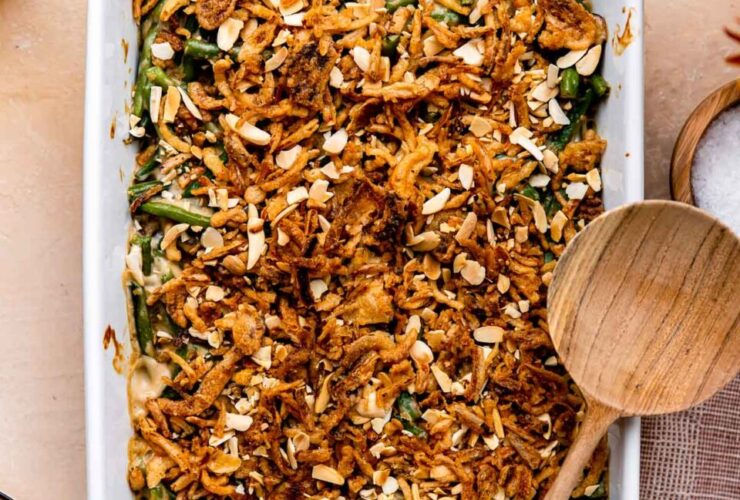A golden-topped green bean casserole in a white ceramic baking dish, garnished with extra crispy fried onions, shot from a slight angle on a rustic wood Thanksgiving table