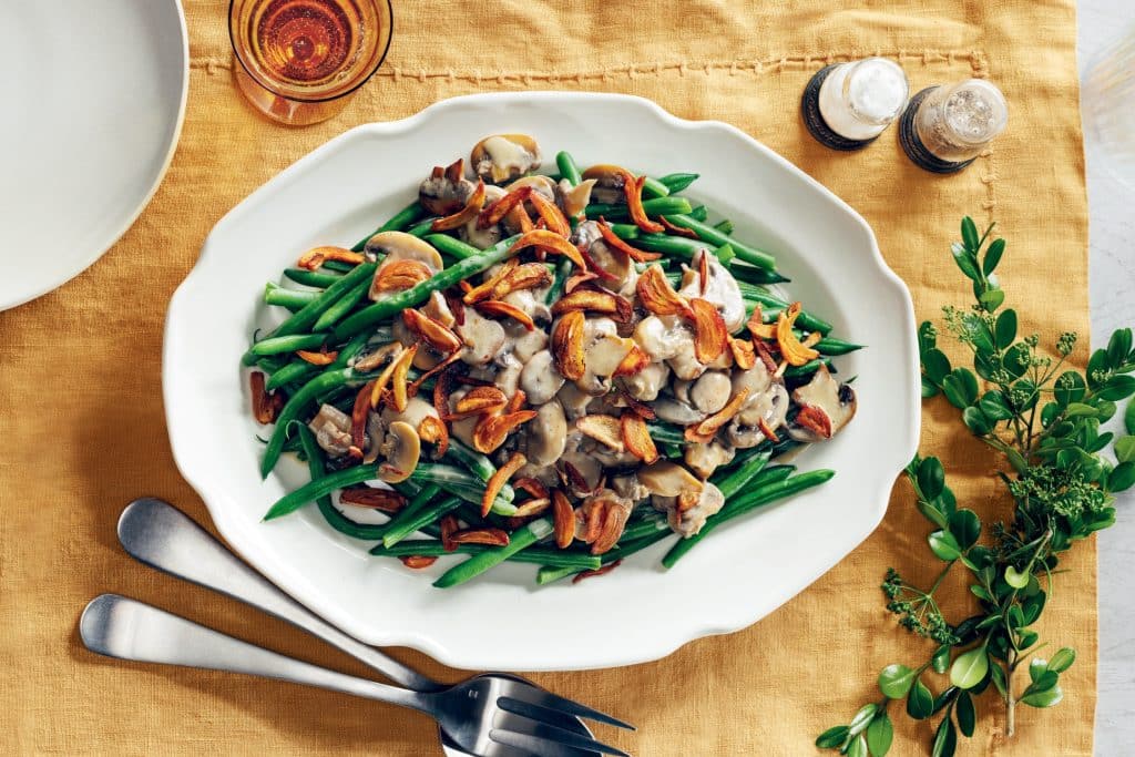 Overhead flat-lay of fresh green beans, sliced cremini mushrooms, a bowl of flour-dredged onion rings, a small pitcher of cream, and shredded Gruyère on a marble surface