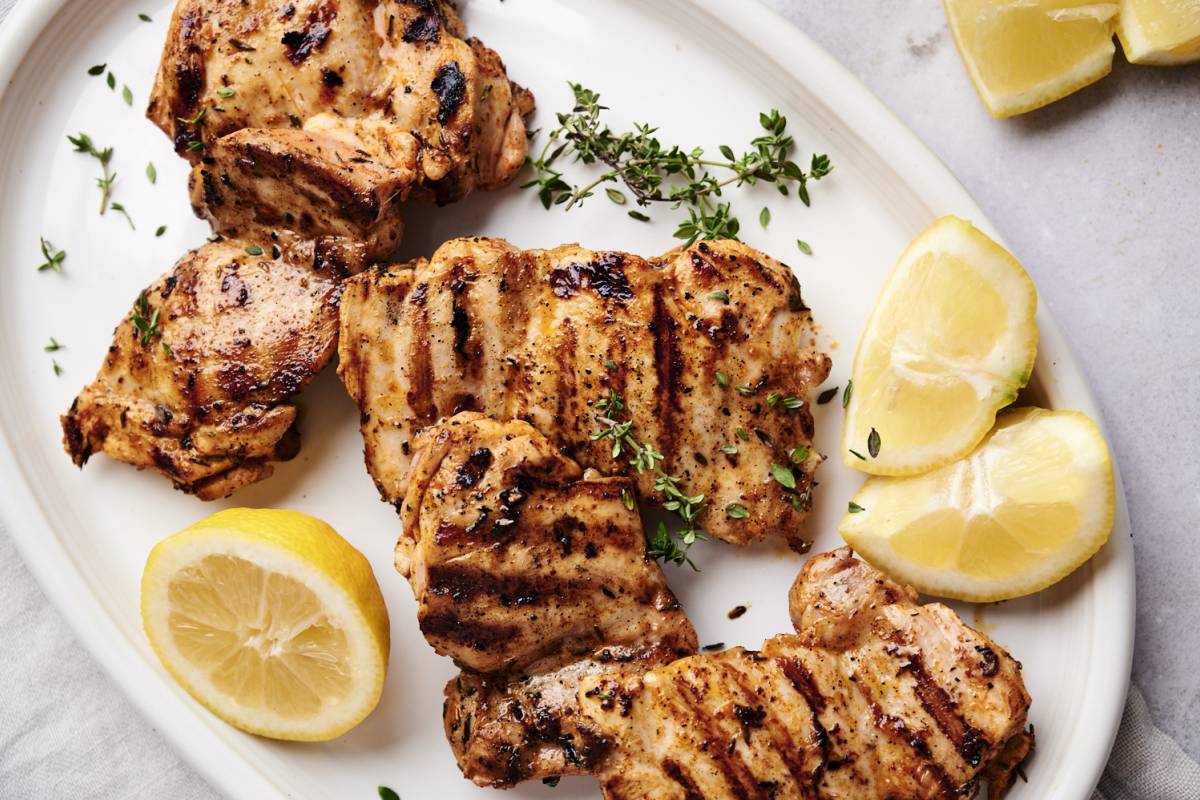 Overhead flat lay of raw chicken thighs in a glass bowl with marinade ingredients — olive oil, garlic, lemon, smoked paprika, and fresh herbs arranged around the bowl
