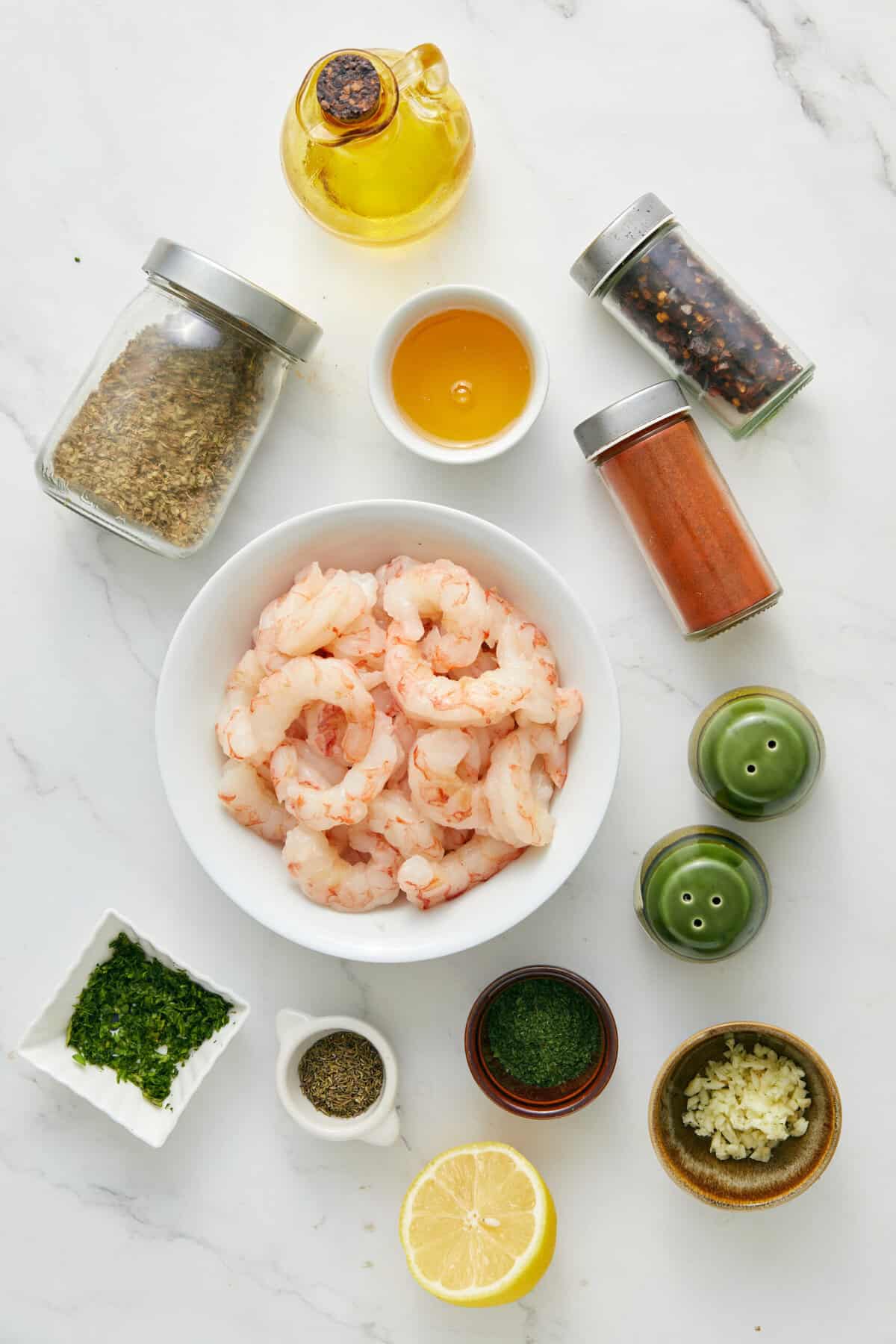 Overhead flat-lay of raw shrimp in a glass bowl with marinade ingredients surrounding it — smoked paprika, garlic, lemon, olive oil, and fresh parsley on a light marble surface