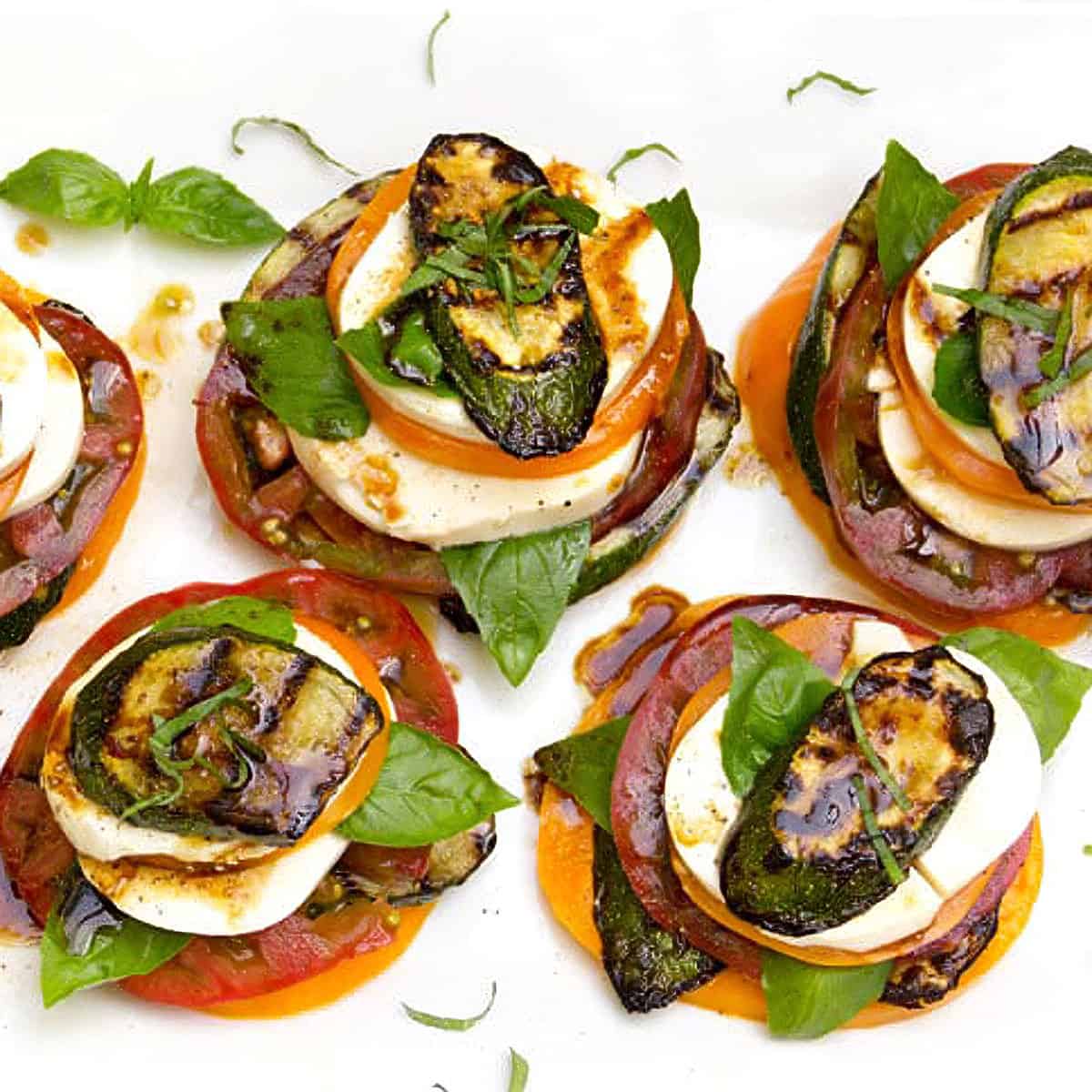 Grilled zucchini caprese stacks arranged on a white marble serving board, drizzled with balsamic glaze and garnished with fresh basil leaves