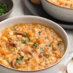 A rustic bowl of ham and bean soup with creamy white beans, tender chunks of ham, and fresh parsley garnish, photographed overhead on a wooden table