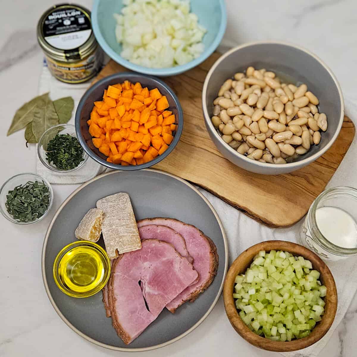 Dried white beans soaking in a bowl of water next to a ham bone, chopped carrots, celery, and onion on a wooden cutting board — ingredients shot for ham and bean soup