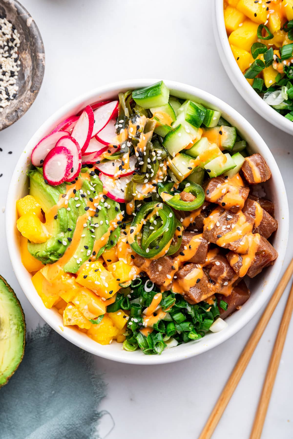 Close-up of a Hawaiian poke bowl showing glistening cubed ahi tuna coated in sesame-soy marinade, topped with sliced avocado, sesame seeds, and a sriracha mayo drizzle, shallow depth of field