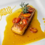 Herb-crusted salmon fillet with golden herb crust served on a white platter surrounded by roasted summer vegetables including zucchini, cherry tomatoes, and bell peppers, garnished with fresh basil and lemon wedges