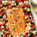 Herb-crusted salmon fillet with golden herb crust served on a white platter surrounded by roasted summer vegetables including zucchini, cherry tomatoes, and bell peppers, garnished with fresh basil and lemon wedges