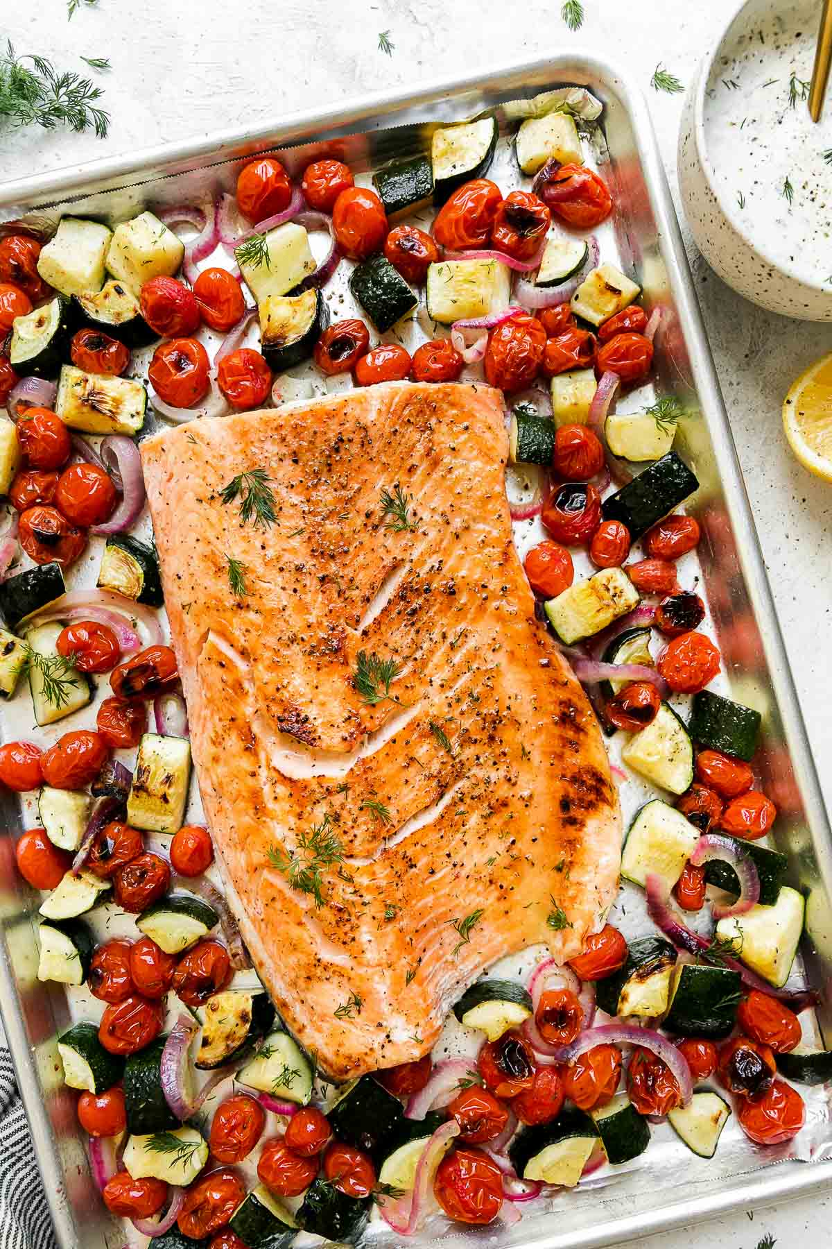Herb-crusted salmon fillet with golden herb crust served on a white platter surrounded by roasted summer vegetables including zucchini, cherry tomatoes, and bell peppers, garnished with fresh basil and lemon wedges