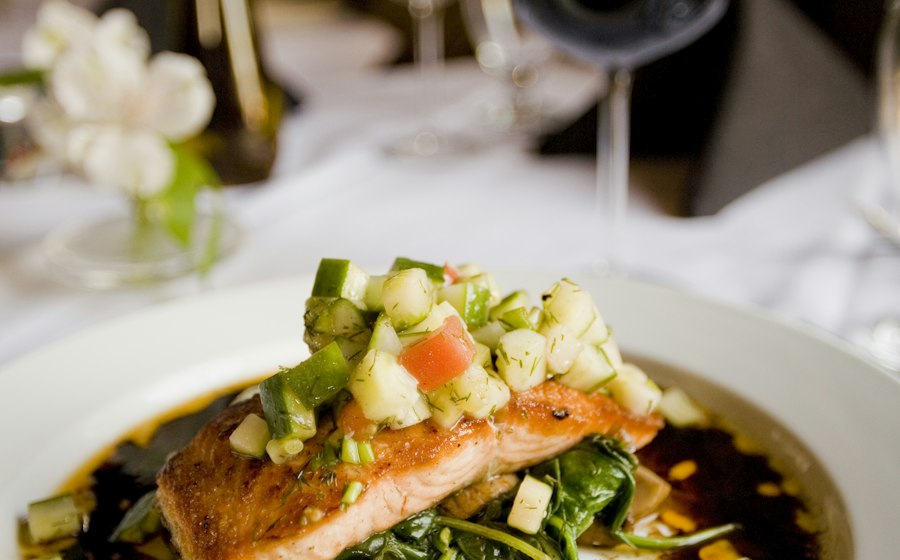 Close-up of herb-crusted salmon fillet with a golden-green parsley and dill crust, resting on a bed of blistered cherry tomatoes and roasted zucchini, with a lemon wedge being squeezed over the top