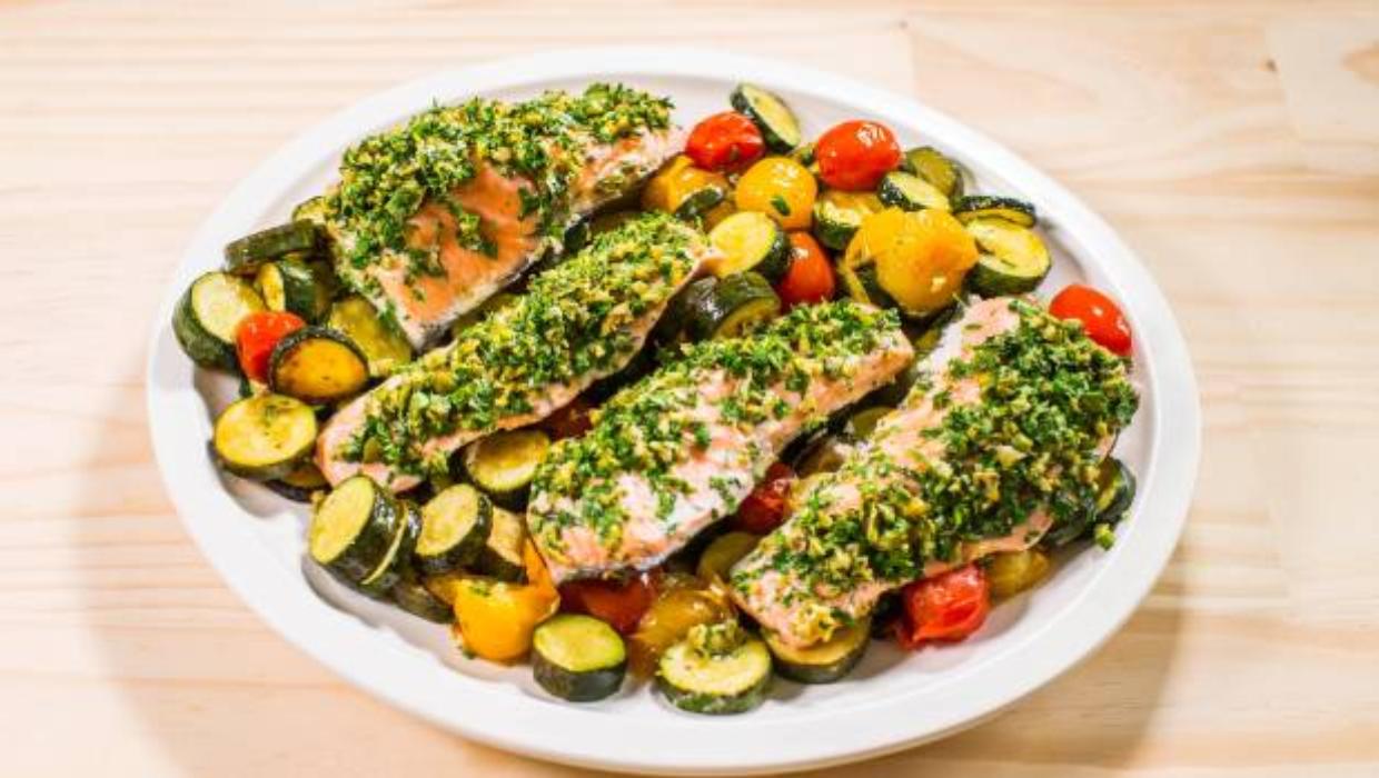 Close-up of herb-crusted salmon fillet with a golden-green parsley and dill crust, resting on a bed of blistered cherry tomatoes and roasted zucchini, with a lemon wedge being squeezed over the top