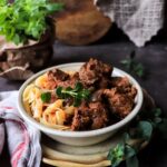 A beautifully plated bowl of Hungarian Goulash with tender beef chunks in a deep red paprika broth, garnished with fresh parsley and served alongside egg noodles, photographed on a rustic wooden table