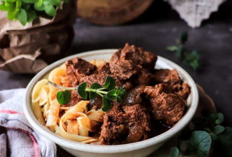 A beautifully plated bowl of Hungarian Goulash with tender beef chunks in a deep red paprika broth, garnished with fresh parsley and served alongside egg noodles, photographed on a rustic wooden table