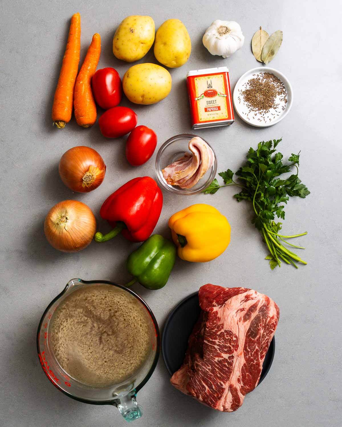 An overhead flat-lay of the key Hungarian Goulash ingredients — chunks of beef chuck, sweet Hungarian paprika, whole onions, garlic cloves, bell peppers, tomato paste, and fresh parsley arranged on a wooden cutting board
