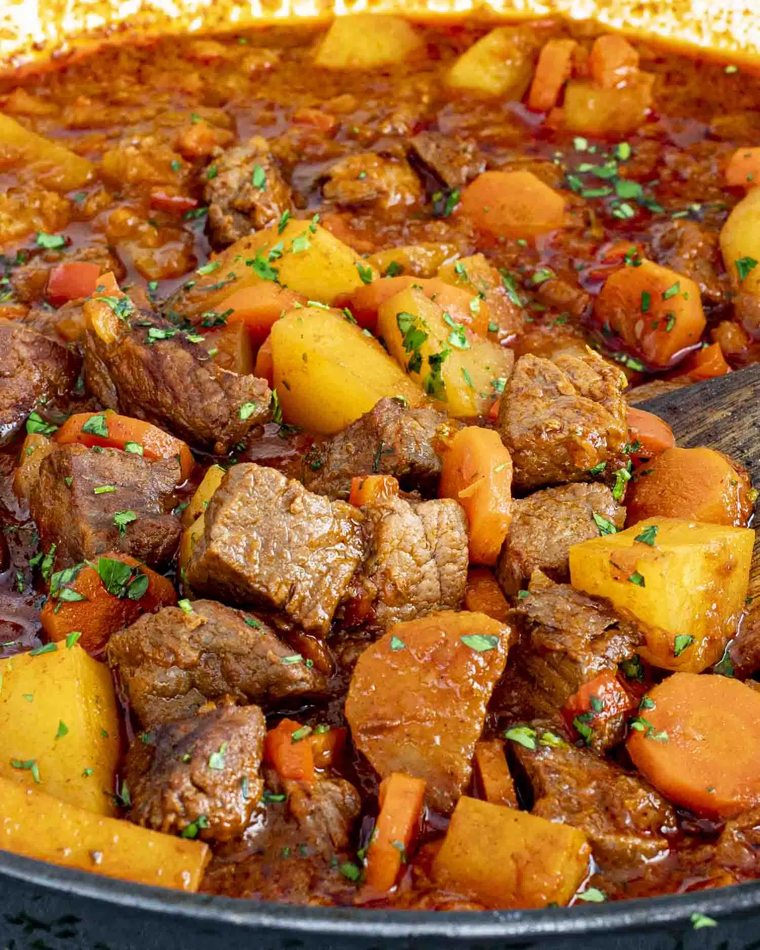 A close-up shot of a ladle lifting Hungarian Goulash from the pot, showing tender beef chunks, glistening red paprika broth, and soft bell pepper pieces, with steam rising and fresh parsley visible