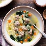 A wide ceramic bowl of Italian wedding soup with golden broth, tender mini meatballs, bright green spinach, and tiny acini de pepe pasta, garnished with freshly grated Parmesan and cracked black pepper, photographed from above on a linen napkin with a rustic wooden spoon