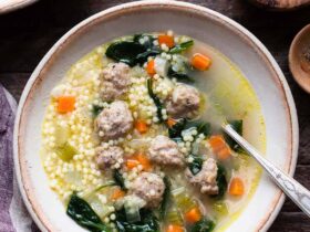 A wide ceramic bowl of Italian wedding soup with golden broth, tender mini meatballs, bright green spinach, and tiny acini de pepe pasta, garnished with freshly grated Parmesan and cracked black pepper, photographed from above on a linen napkin with a rustic wooden spoon