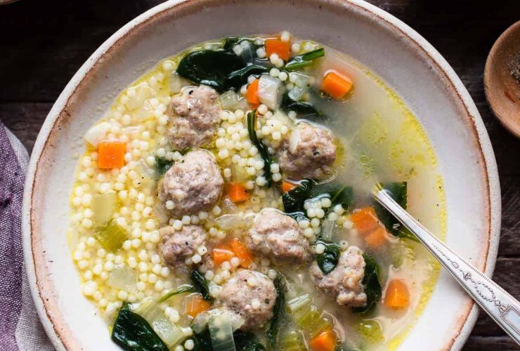 A wide ceramic bowl of Italian wedding soup with golden broth, tender mini meatballs, bright green spinach, and tiny acini de pepe pasta, garnished with freshly grated Parmesan and cracked black pepper, photographed from above on a linen napkin with a rustic wooden spoon