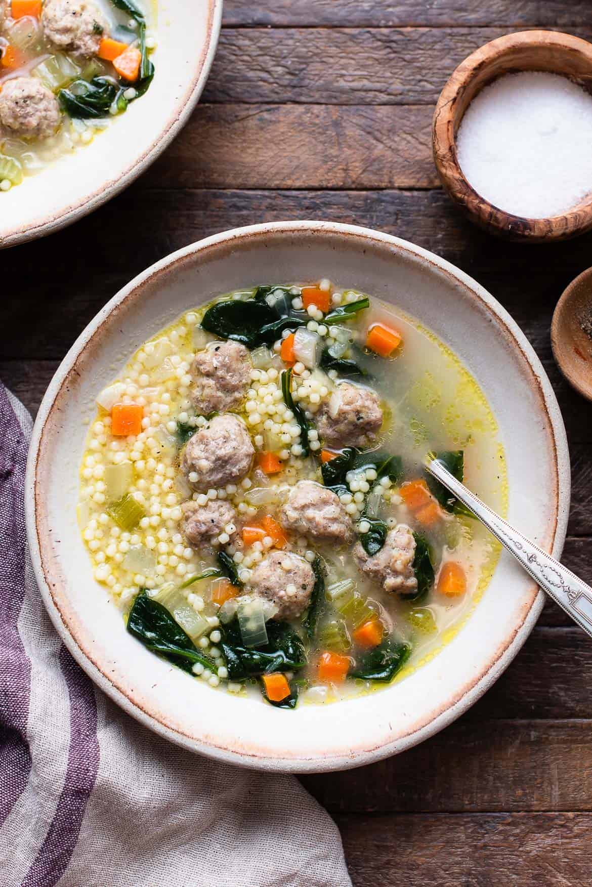 A wide ceramic bowl of Italian wedding soup with golden broth, tender mini meatballs, bright green spinach, and tiny acini de pepe pasta, garnished with freshly grated Parmesan and cracked black pepper, photographed from above on a linen napkin with a rustic wooden spoon