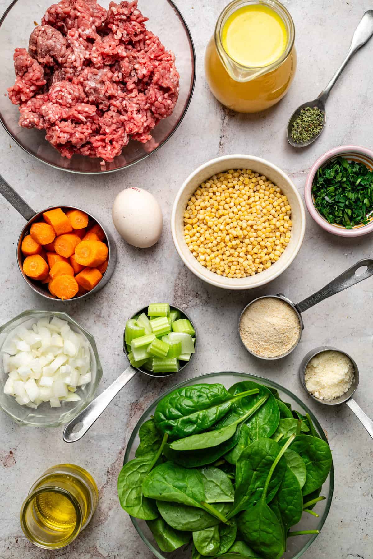 A flat lay of Italian wedding soup ingredients including a bowl of ground meat mixture, a pile of fresh baby spinach, a small bowl of acini de pepe pasta, a container of chicken broth, minced garlic, fresh parsley, and grated Parmesan on a marble surface