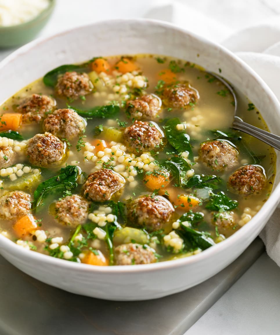A close-up of a ladle lifting Italian wedding soup out of a large Dutch oven, showing glistening golden broth, perfectly round mini meatballs, bright wilted spinach, and tiny acini de pepe pasta, steam rising gently in soft kitchen light