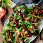 Overhead shot of healthy Korean BBQ lettuce wrap bowls styled on a wooden board with marinated grilled beef, crisp butter lettuce cups, shredded carrots, cucumbers, and sesame seeds, with small dipping sauce bowls on the side
