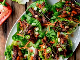 Overhead shot of healthy Korean BBQ lettuce wrap bowls styled on a wooden board with marinated grilled beef, crisp butter lettuce cups, shredded carrots, cucumbers, and sesame seeds, with small dipping sauce bowls on the side