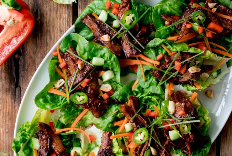 Overhead shot of healthy Korean BBQ lettuce wrap bowls styled on a wooden board with marinated grilled beef, crisp butter lettuce cups, shredded carrots, cucumbers, and sesame seeds, with small dipping sauce bowls on the side