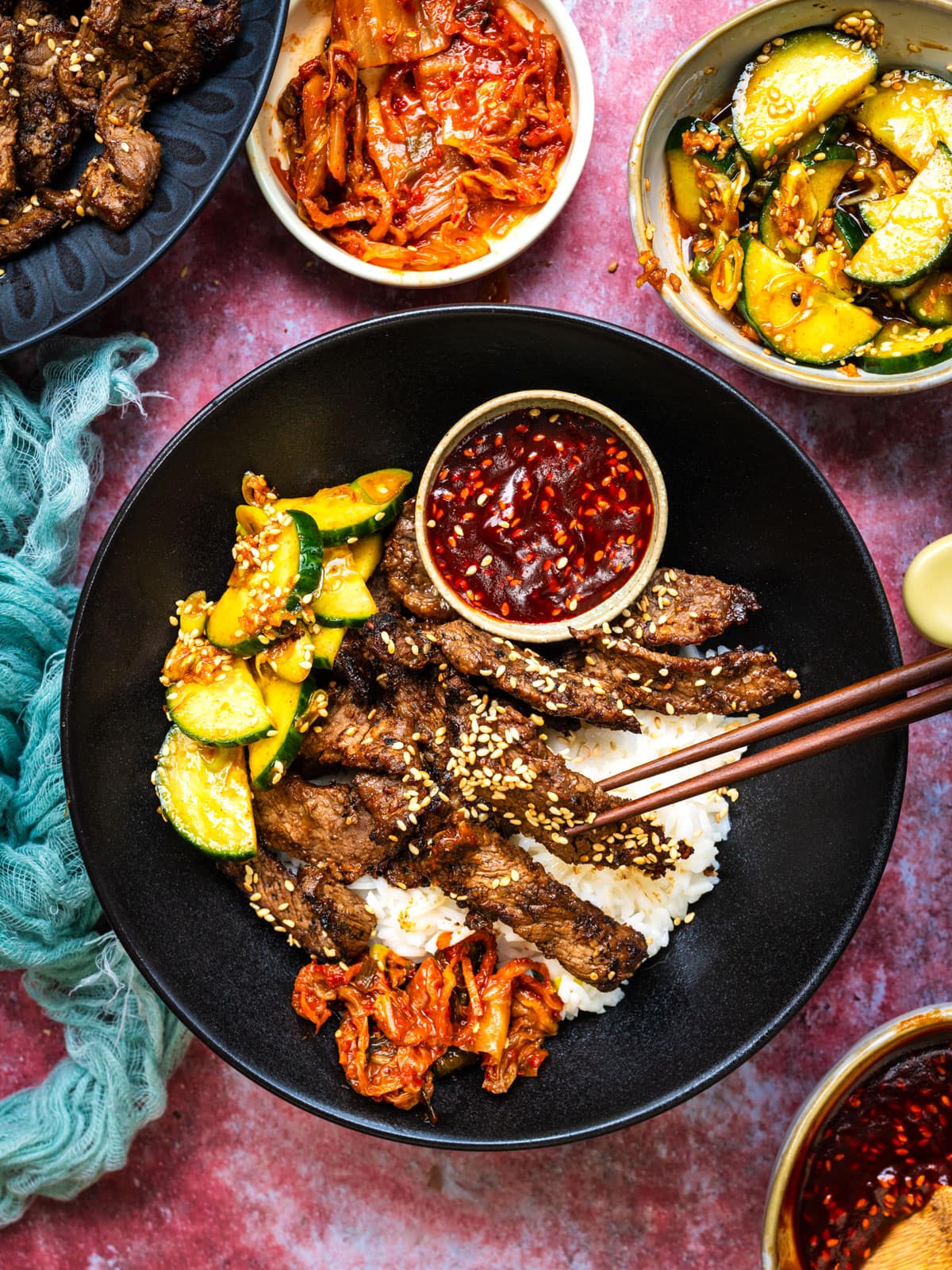 Flat lay ingredients shot showing thinly sliced beef in marinade, fresh garlic, ginger root, sesame oil, soy sauce, butter lettuce leaves, shredded carrots, sliced cucumbers, and small bowls of gochujang and sesame seeds arranged on a light marble surface