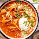 A rustic ceramic bowl filled with lasagna soup topped with a swirl of creamy ricotta, shredded mozzarella, and fresh basil, photographed in warm natural light on a dark wood surface
