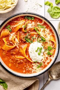 A rustic ceramic bowl filled with lasagna soup topped with a swirl of creamy ricotta, shredded mozzarella, and fresh basil, photographed in warm natural light on a dark wood surface