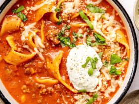 A rustic ceramic bowl filled with lasagna soup topped with a swirl of creamy ricotta, shredded mozzarella, and fresh basil, photographed in warm natural light on a dark wood surface