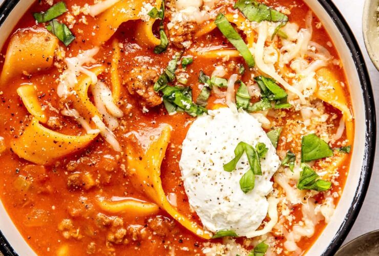 A rustic ceramic bowl filled with lasagna soup topped with a swirl of creamy ricotta, shredded mozzarella, and fresh basil, photographed in warm natural light on a dark wood surface