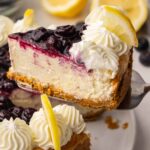 A beautifully plated slice of lemon blueberry cheesecake on a white plate with blueberry sauce drizzled over the top and curled lemon slices as garnish, shot in natural light