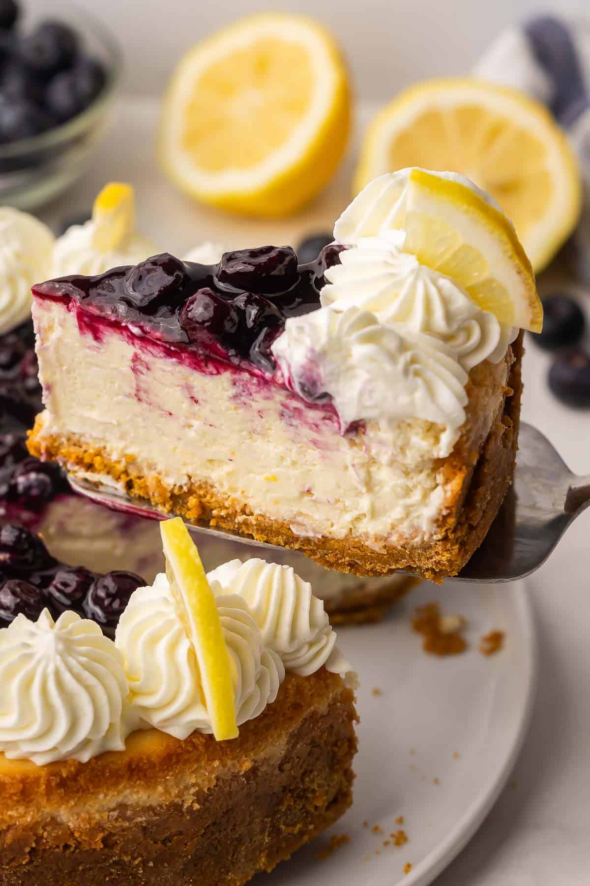 A beautifully plated slice of lemon blueberry cheesecake on a white plate with blueberry sauce drizzled over the top and curled lemon slices as garnish, shot in natural light