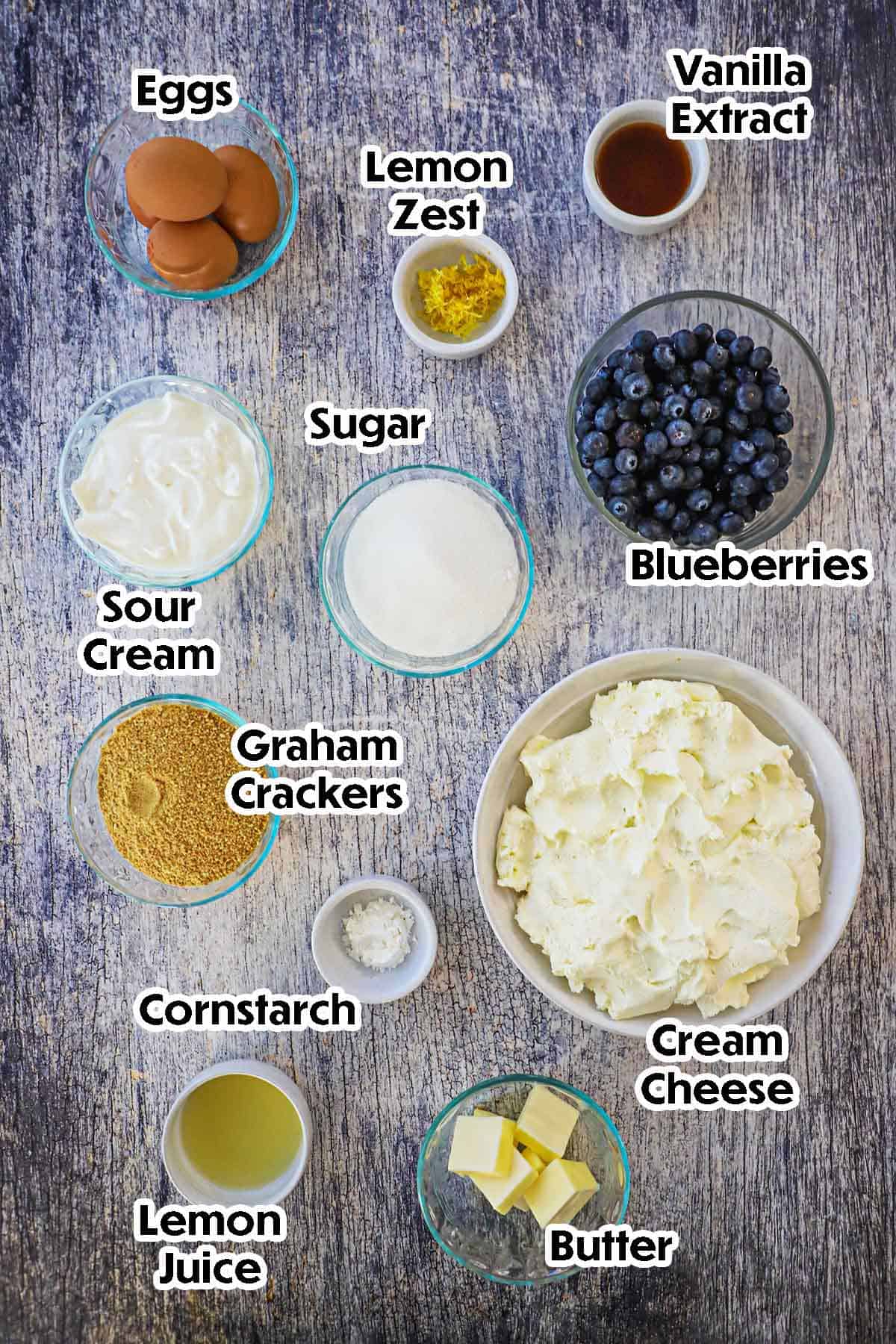 Overhead flat lay of lemon blueberry cheesecake ingredients — cream cheese blocks, fresh lemons, blueberries, breadcrumbs, butter, eggs, and sugar on a marble surface