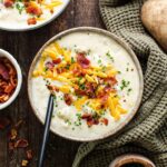 A steaming bowl of loaded baked potato soup topped with crispy bacon crumbles, shredded cheddar cheese, sour cream, and fresh chives, photographed on a dark wood surface with a rustic linen napkin