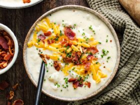 A steaming bowl of loaded baked potato soup topped with crispy bacon crumbles, shredded cheddar cheese, sour cream, and fresh chives, photographed on a dark wood surface with a rustic linen napkin
