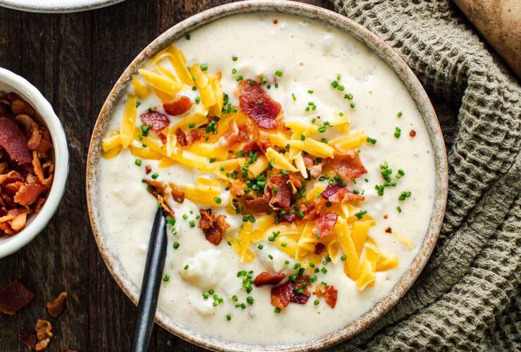 A steaming bowl of loaded baked potato soup topped with crispy bacon crumbles, shredded cheddar cheese, sour cream, and fresh chives, photographed on a dark wood surface with a rustic linen napkin