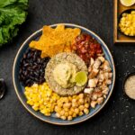 Mexican street corn quinoa salad served in a wide white bowl, garnished with cotija cheese, fresh cilantro, chili powder, and lime wedges on a bright wooden surface