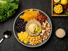 Mexican street corn quinoa salad served in a wide white bowl, garnished with cotija cheese, fresh cilantro, chili powder, and lime wedges on a bright wooden surface