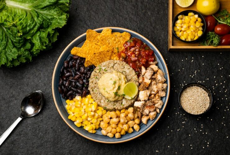 Mexican street corn quinoa salad served in a wide white bowl, garnished with cotija cheese, fresh cilantro, chili powder, and lime wedges on a bright wooden surface