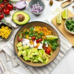 Mexican street corn quinoa salad served in a wide white bowl, garnished with cotija cheese, fresh cilantro, chili powder, and lime wedges on a bright wooden surface