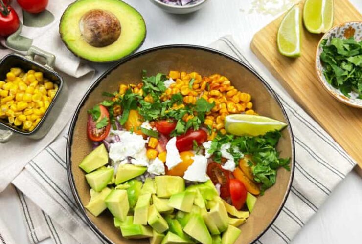 Mexican street corn quinoa salad served in a wide white bowl, garnished with cotija cheese, fresh cilantro, chili powder, and lime wedges on a bright wooden surface