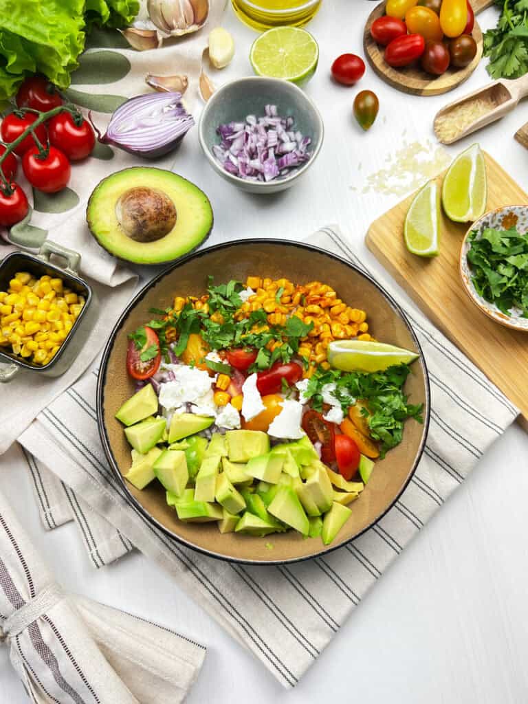Mexican street corn quinoa salad served in a wide white bowl, garnished with cotija cheese, fresh cilantro, chili powder, and lime wedges on a bright wooden surface