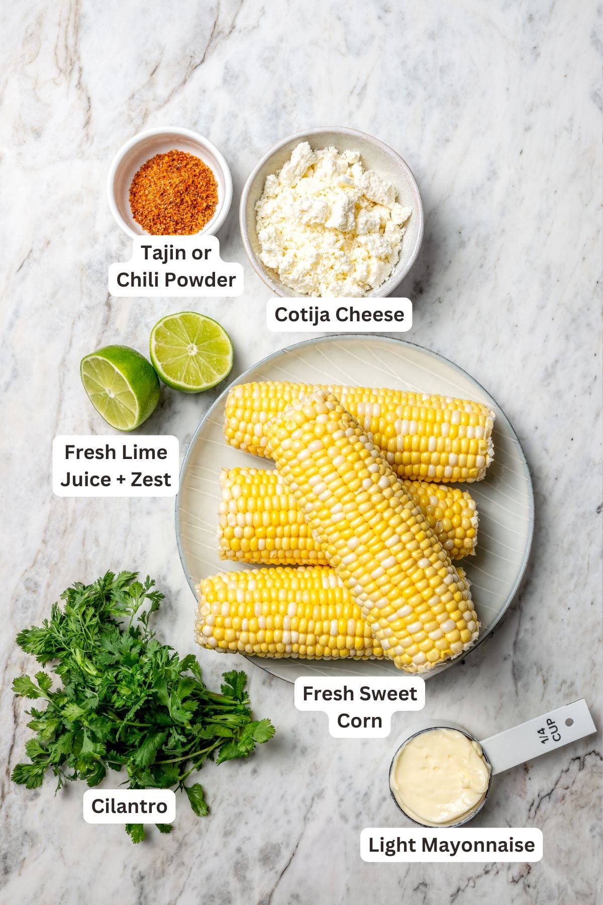Overhead flat lay of Mexican street corn quinoa salad ingredients — quinoa, corn, lime, cotija, cilantro, jalapeño, chili powder, and red onion arranged on a marble surface