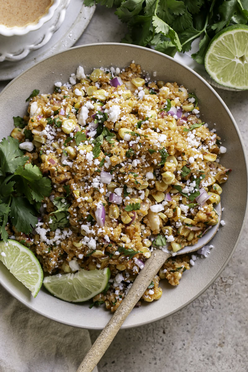 Close-up of Mexican street corn quinoa salad in a serving bowl with visible cotija crumbles, fresh cilantro leaves, charred corn kernels, and a halved lime on the side